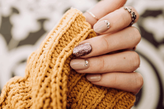 Es ist eine Frauenhand mit steinernen und glitzernen Fingernägeln an zu sehen. Die Frau hat einen dunkelgelben Pullover an.
