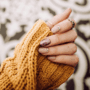 Es ist eine Frauenhand mit steinernen und glitzernen Fingernägeln an zu sehen. Die Frau hat einen dunkelgelben Pullover an.